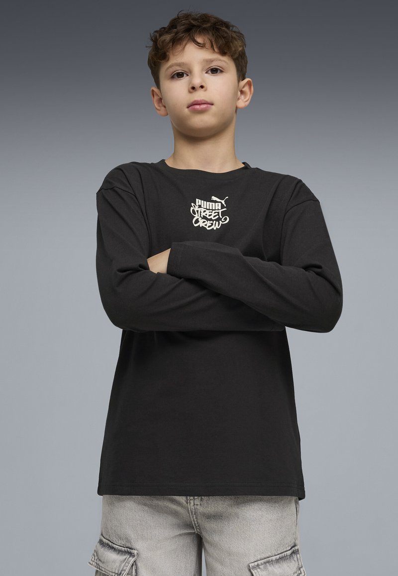 Junge mit verschränkten Armen, der ein schwarzes Puma Street Crew Langarmshirt und hellgraue Cargo-Hosen trägt, vor grauem Hintergrund.