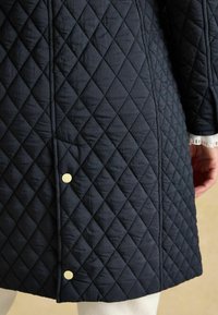 Prešívaný námorní kabát s diamantovým vzorom, ktorý má zlaté cvoky a zvýrazňuje textúru a tvar na leme a rukávoch.