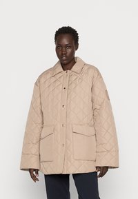 Casaco acolchoado bege com grandes bolsos, fecho de botões e colarinho. Apresenta um design texturizado e almofadado, e um logo cosido na manga.