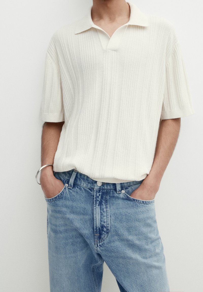 Homme portant un polo en maille côtelée blanche à manches courtes et un jean en denim bleu clair, les mains dans les poches sur fond blanc.