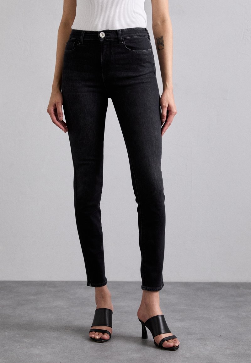 Jeans skinny de mezclilla negra con cintura alta, que cuentan con bolsillos frontales y cierre de botón. Combinados con sandalias de tacón negras.