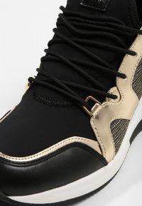 Baskets noires avec des détails beige et or métallique, lacets noirs et semelle blanche, gros plan sur l'orteil et le côté.