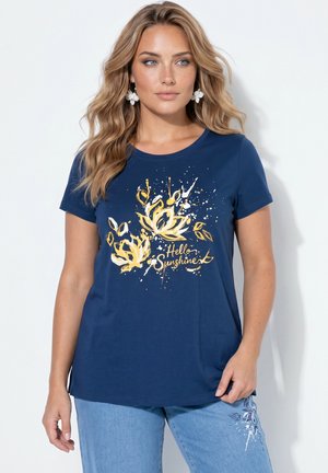 Vrouw met lang blond haar die een blauw T-shirt draagt met een geel bloemenpatroon en tekst "Hello Sunshine" en lichtblauwe spijkerbroek met bloemendetails.