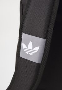 Détail d'un sac à dos en tissu noir avec une étiquette grise arborant un logo à trois bandes blanches et un motif de feuille, mettant en valeur le contraste des matériaux et des couleurs.