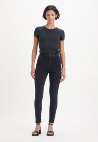 Svart tettsittende t-skjorte med korte ermer og knyting i livet, kombinert med høyhalkede mørke jeans og svarte ankelrem-sandaler.
