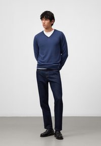 Blauer V-Ausschnitt-Pullover aus Strickstoff, kombiniert mit dunklen Jeans. Modell trägt schwarze Schuhe. Einfaches Design, lockere Passform, glatte Textur.