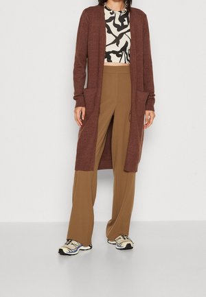 Femme portant un long cardigan marron, un crop top à motifs noirs et blancs, un pantalon large marron taille haute, et des baskets beige.