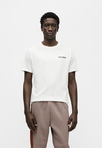 Λευκό βαμβακερό μπλουζάκι με πράσινο λογότυπο "Converse" στο στήθος, συνδυασμένο με παντελόνια καρό μοτίβο σε καφέ, πορτοκαλί και μπεζ αποχρώσεις.