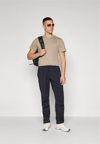 Bež kratka majica s kratkimi rokavi, temnomodra elastika za pas hlače, sive superge, sončna očala in črn nahrbtnik. Nevtralna barvna paleta in priložnostna zasnova.