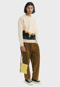 Giovane con i capelli ricci che indossa un maglione beige con motivo arancione scuro e nero, pantaloni in velluto a coste marroni, sneakers nere, mentre tiene una borsa gialla.