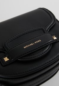 Sac à main en cuir noir avec une texture lisse, une forme arrondie, des accents en métal doré et un emblème signature de Michael Kors.