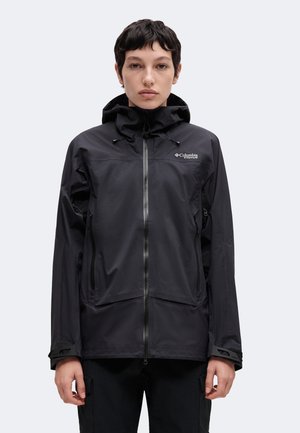 Personne portant une veste imperméable noire Columbia Titanium avec capuche, debout face à l’avant, expression neutre, fond clair uni.