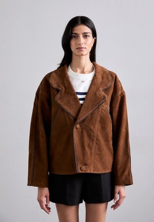 Donna con lunghi capelli scuri che indossa una giacca oversize in suede marrone sopra una camicia bianca e pantaloni corti neri, in piedi di fronte a uno sfondo grigio uniforme.
