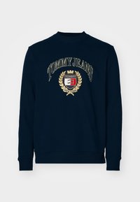 Sudadera azul marino con cuello redondo, que presenta un logotipo de Tommy Jeans bordado en dorado y un escudo circular con un diseño de corona y laurel.