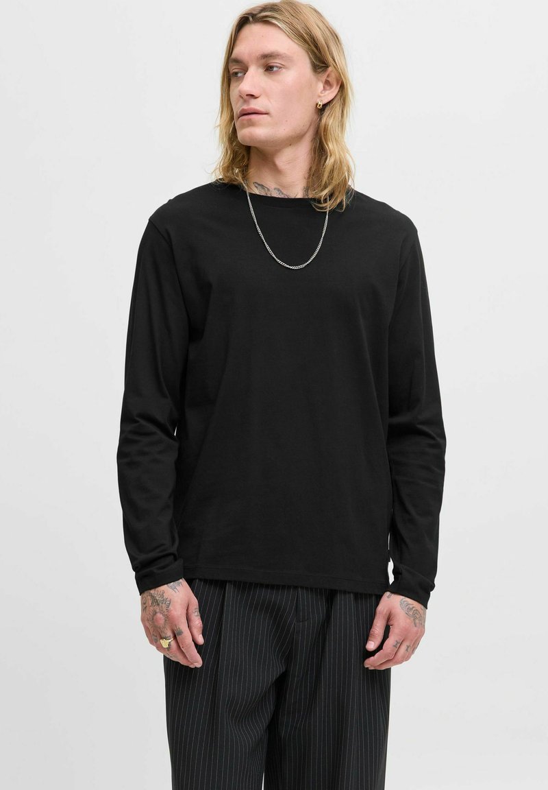 Schwarz langärmliges T-Shirt mit Rundhalsausschnitt, aus weichem Stoff gefertigt. Trägt zu einer gestreiften Hose. Zusätzliche Details umfassen eine silberne Kette.