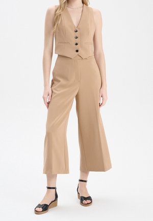 Beżowa krótka kamizelka z pięcioma czarnymi guzikami i dwiema kieszeniami, zestawiona z wysokim stanem szerokimi spodniami. Czarne sandały na koturnie z paskami dopełniają całość.