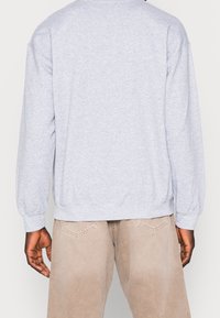 Sudadera gris claro de manga larga, con puños y dobladillo acanalados, fabricada en una tela suave. Llevada con pantalones beige, mostrando un ajuste relajado en la parte de atrás.