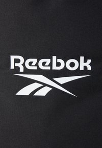 Čierna tkanina s veľkým bielym logom Reebok, so štýlovým textom a zakrivenými líniami, ktoré zdôrazňujú dizajnovú estetiku značky.