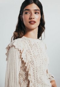 Giovane donna con lunghi capelli castano ondulati indossa un maglione lavorato a maglia color crema con rifiniture in pizzo, guardando di lato su uno sfondo semplice.