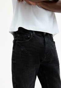 Person iført sorte slidte jeans og en hvid skjorte med hænderne foldet over brystet foran en hvid baggrund.
