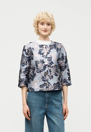 Νεαρό άτομο που φοράει ένα μπλε σακάκι με φλοράλ μοτίβο, κουμπωτό, με μανίκια τριών τετάρτων και ανοιχτογάλατσινο τζιν, μπροστά σε απλό φόντο.