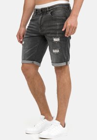 Shorts de mezclilla gris oscuro con detalles desgastados y dobladillos deshilachados, con cinco bolsillos y un ajuste ligeramente relajado. Combinados con zapatillas blancas.