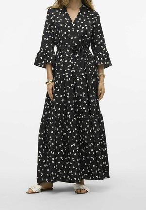 Femme portant une robe noire longueur cheville avec des motifs en forme de cœur blancs, des manches cloche et une ceinture à la taille, associée à des sandales blanches à bout ouvert.