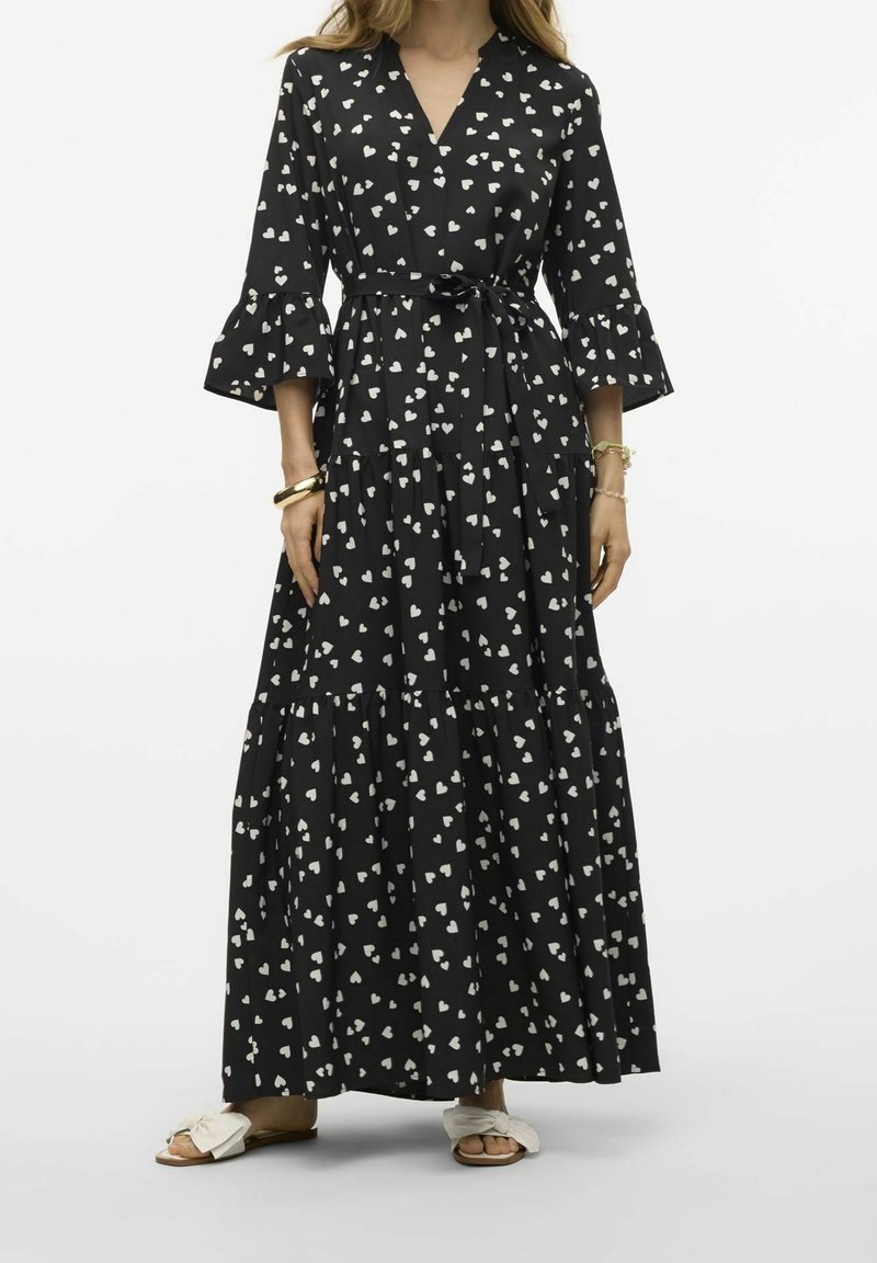 Femme portant une robe noire longueur cheville avec des motifs en forme de cœur blancs, des manches cloche et une ceinture à la taille, associée à des sandales blanches à bout ouvert.