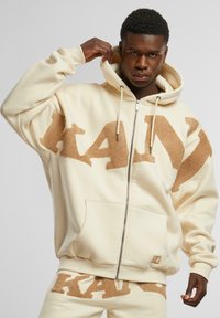 Kremfarget hettegenser med glidelås, stor beige "KANY" logo, forlommer og snor. Laget av myk, teksturert stoff.