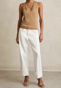 Brązowy, bezrękawnik z dekoltem w serek o połyskującej fakturze, w połączeniu z białymi, wysokim stanem, podwiniętymi dżinsami oraz beżowymi sandałami na słupku.