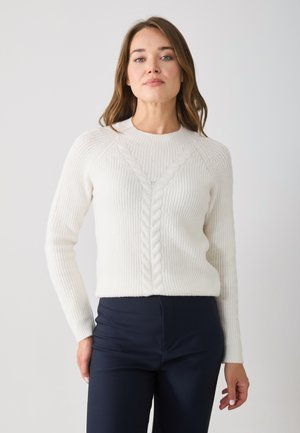 Pull en maille blanche à texture côtelée, avec un design en câble en forme de V. Associé à un pantalon ajusté bleu marine, présenté sur un fond neutre.