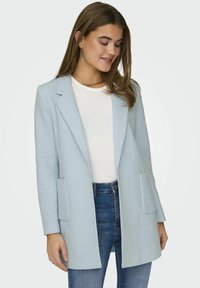Bluza lungă de culoare albastră deschisă, cu o finisare texturată, rever în formă de v și două buzunare frontale, asortată cu un top alb și blugi.