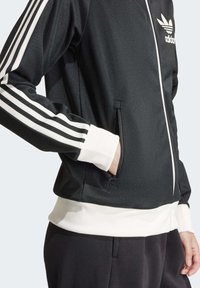 Černá sportovní bunda s bílými proužky na rukávech, kapsami na zip po stranách a žebrovanými manžetami; na hrudi je logo Adidas.