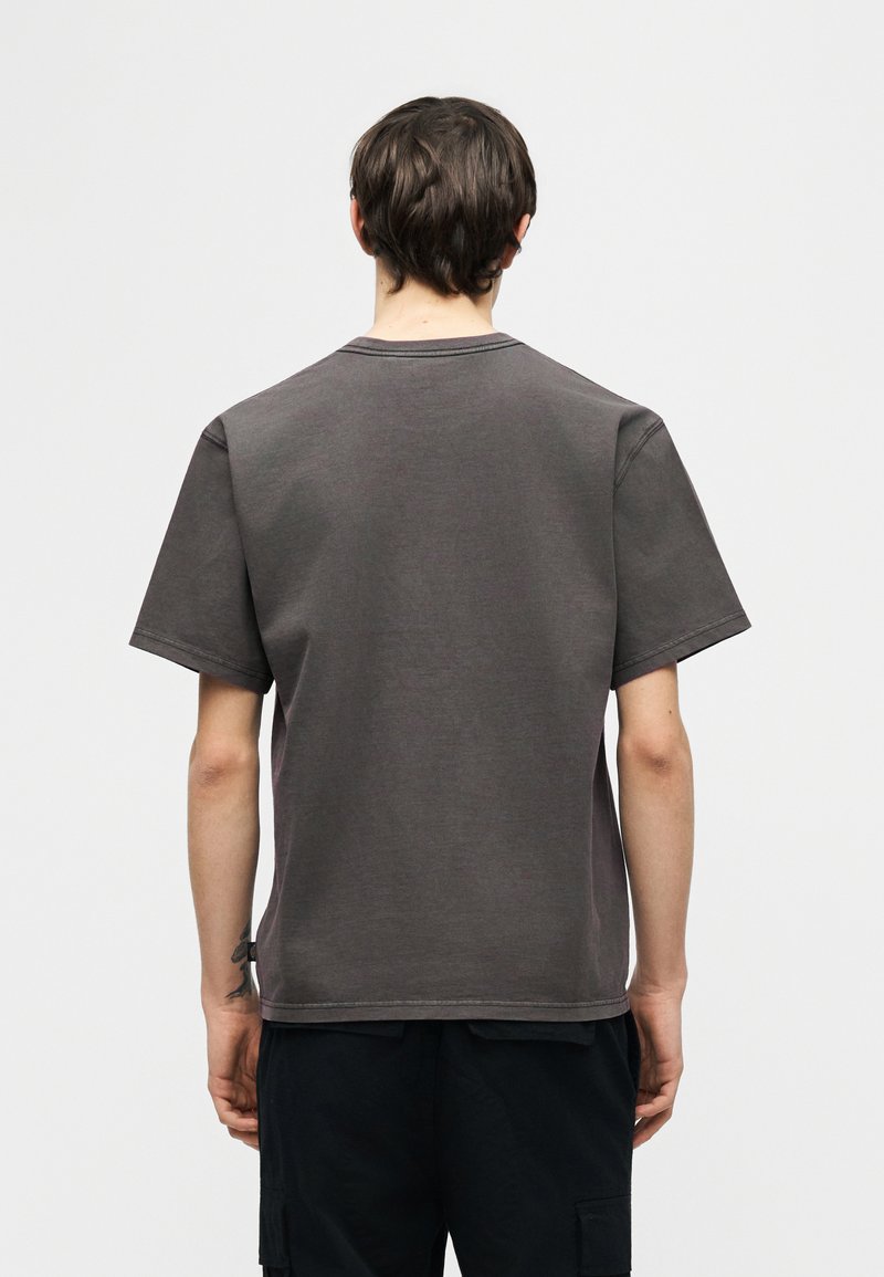 Giovane in piedi rivolto di spalle, indossa una t-shirt a maniche corte grigio scuro semplice e pantaloni neri su uno sfondo bianco.