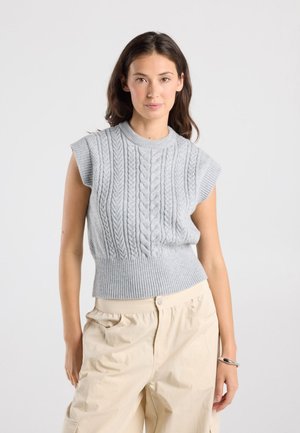 Jonge vrouw draagt lichtgrijze mouwloze kabelgebreide trui en beige broek, staat met één hand in de zak tegen een effen achtergrond.