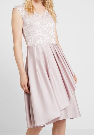 Ärmelloses hellrosa Kleid mit Spitzenblumenoberteil und glänzendem plissiertem Satinstoffrock, der am Knie endet, getragen von einer Frau mit blondem Haar.