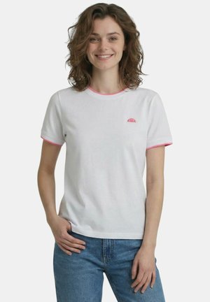 Giovane donna sorridente che indossa una maglietta bianca con bordino rosa e jeans blu, in piedi con una mano in tasca su sfondo bianco.