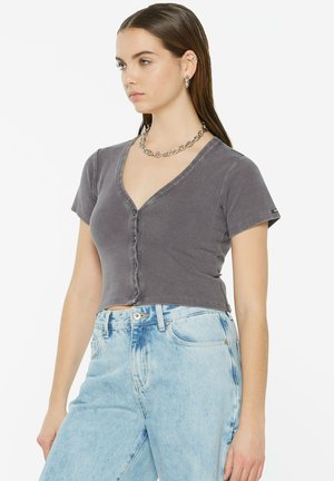 Graues, kurzärmliges, bauchfreies Button-Up-T-Shirt mit V-Ausschnitt, kombiniert mit hellblauen, hoch taillierten Jeans. Sichtbare silberne Kette.