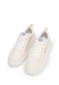 Zapatillas de lona beige con cordones blancos, suela de goma gruesa y texturizada, y una etiqueta con el logo en la lengüeta. Presentan una tira en el talón.