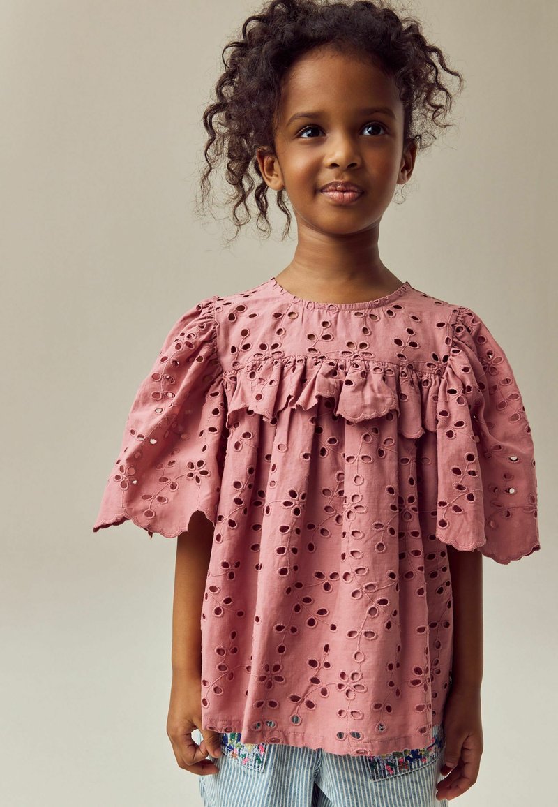 Rosa kurzärmelig Bluse aus Broderie anglaise Stoff, mit floralen Mustern, Rüschendetail am Halsauschnitt und gewellten Kanten.