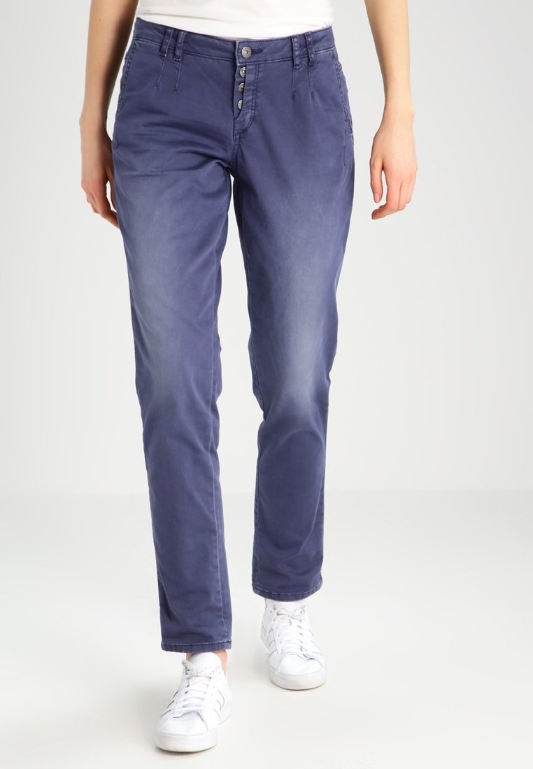 Vrouw in blauwe rechte broek met zichtbare knopen en witte sneakers, staande voor een effen lichte achtergrond.