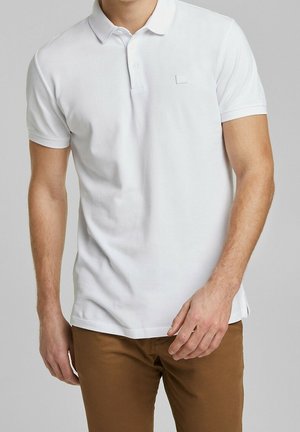 Mann trägt ein schlichtes weißes Polohemd mit kurzen Ärmeln und braune Hose, steht vor einem neutralgrauen Hintergrund.