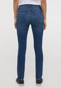 Tmavomodré džínsy so slim fit strihom, klasickým dizajnom s piatimi vreckami a unikátnym prešívaním zadného vrecka. Nosené s čiernymi teniskami.