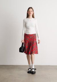 Haut à manches longues blanc, jupe évasée à carreaux rouges, sac à main noir, chaussettes blanches, chaussures Mary Jane noires avec des sangles, debout sur un sol gris.