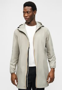 Νεαρός άνδρας που φοράει μπεζ ανοιχτόχρωμο αδιάβροχο με κουκούλα πάνω από ένα λευκό πουκάμισο και μαύρο παντελόνι, κοιτάζει προς τα αριστερά του σε απλό φόντο.