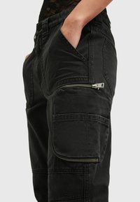 Zwarte cargobroek gemaakt van duurzaam materiaal. Kenmerken zijn onder andere zijzakken en een zichtbare metalen rits op de dij. Gelaagd, casual ontwerp.