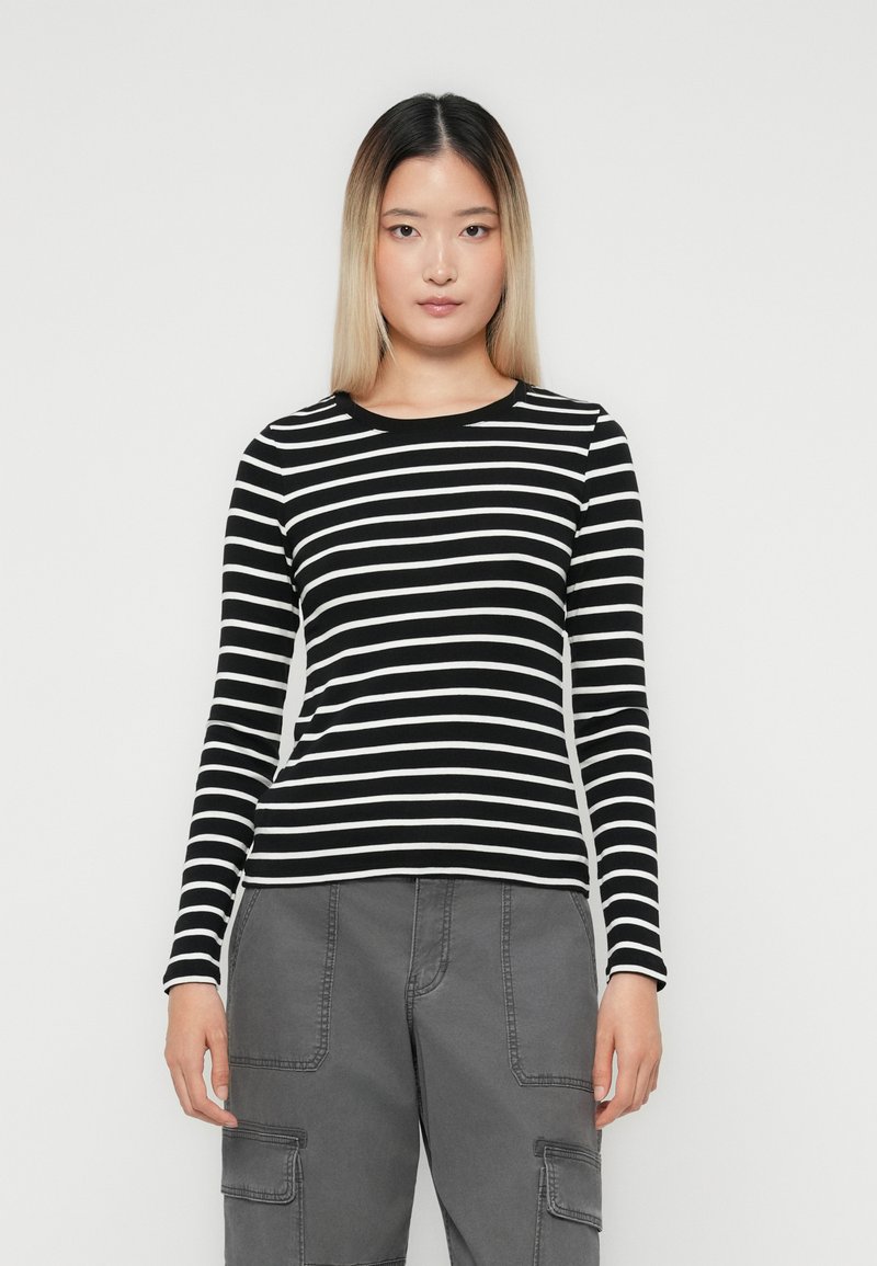 Woman with straight ombre hair wearing a black and white striped long-sleeve shirt and gray cargo pants, standing against a plain background.