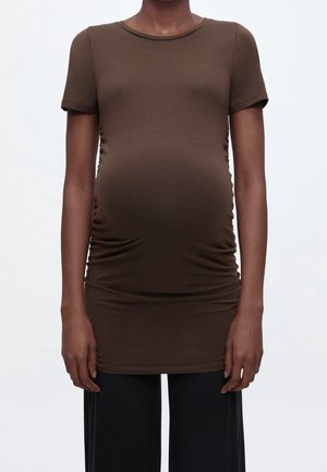 Person iført en tætsiddende brun kortærmet top, der er udstrakt over en gravid mave, kombineret med løse sorte bukser mod en ensfarvet baggrund.