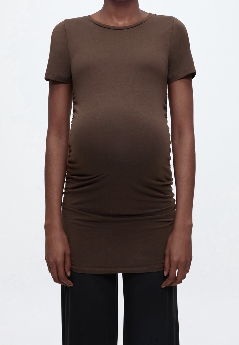 Personne portant un haut marron ajusté à manches courtes tendu sur un ventre de femme enceinte, associé à un pantalon noir ample sur un fond uni.