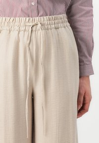 Béžové, lehké kalhoty s uvolněným střihem a elastickým pasem. Obsahují stahovací šňůrku a texturovanou látku. Nad nimi je viditelná pruhovaná košile.